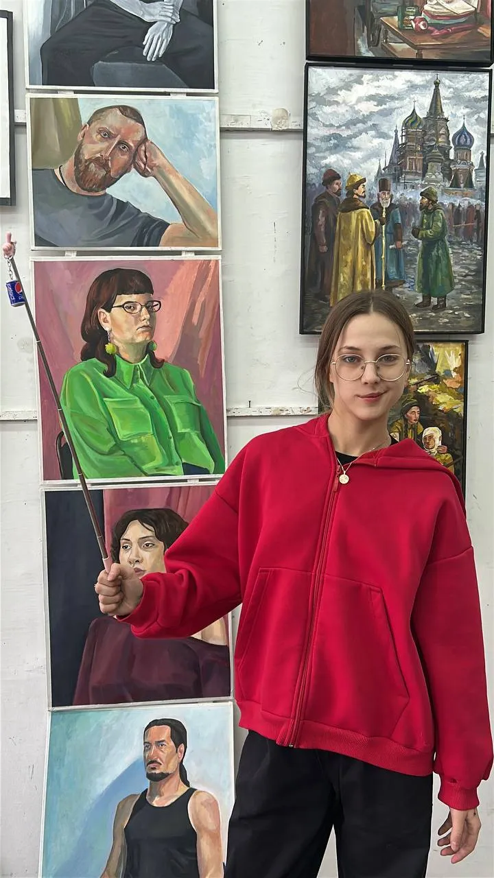 Оля Кубеева в красной толстовке стоит в мастерской на фоне нескольких своих картин